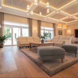 PMP KONKRET SPÓŁKA Z OGRANICZONĄ ODPOWIEDZIALNOŚCIĄ - Jasny salon z drewnianą podłogą, beżowymi sofami i kominkiem z cegły. Sufit z drewnianymi panelami i oświetleniem. Widok na ogród przez duże okna.