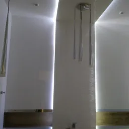 Nowoczesna łazienka z podświetlanym sufitem i ścianami, minimalistyczna lampa wisząca z trzema srebrnymi tubami, widoczna armatura prysznicowa i fragment drewnianego elementu dekoracyjnego.