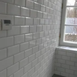 Ściana wyłożona błyszczącymi, białymi płytkami typu metro, widok z boku, obok okno z białymi ramami, na ścianie widoczny panel sterowania