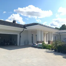 Parterowy dom z garażem, wykończony w jasnych kolorach, z kolumnami przy wejściu, widoczne worki z materiałami budowlanymi i sadzonki roślin na kostce brukowej.