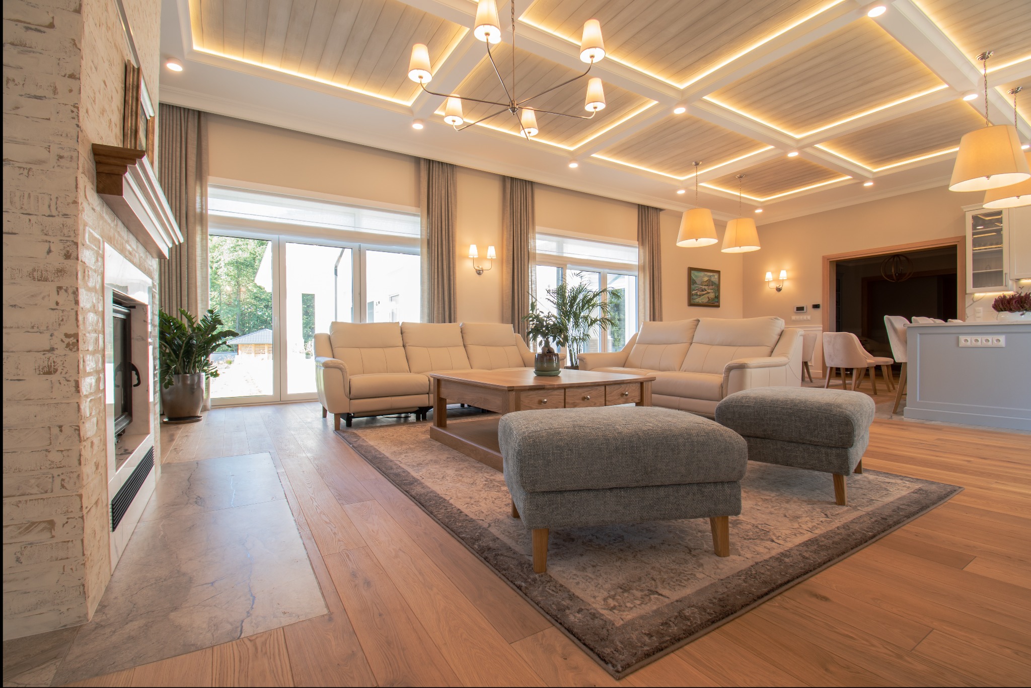 Jasny salon z drewnianą podłogą, beżowymi sofami i kominkiem z cegły. Sufit z drewnianymi panelami i oświetleniem. Widok na ogród przez duże okna.