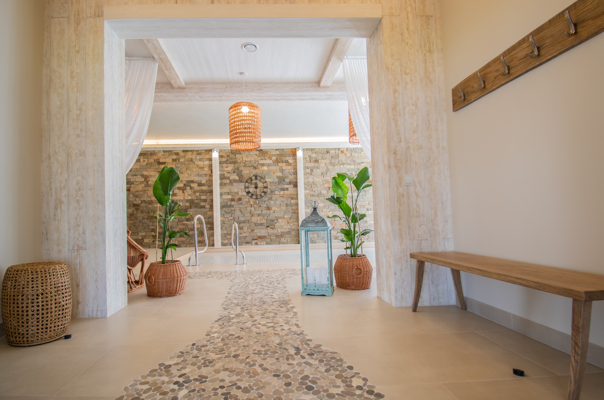 Jasne wnętrze z basenem w tle, dekoracyjne wykończenie podłogi z kamyków, rośliny w wiklinowych donicach, drewniana ławka i wieszak na ścianie.