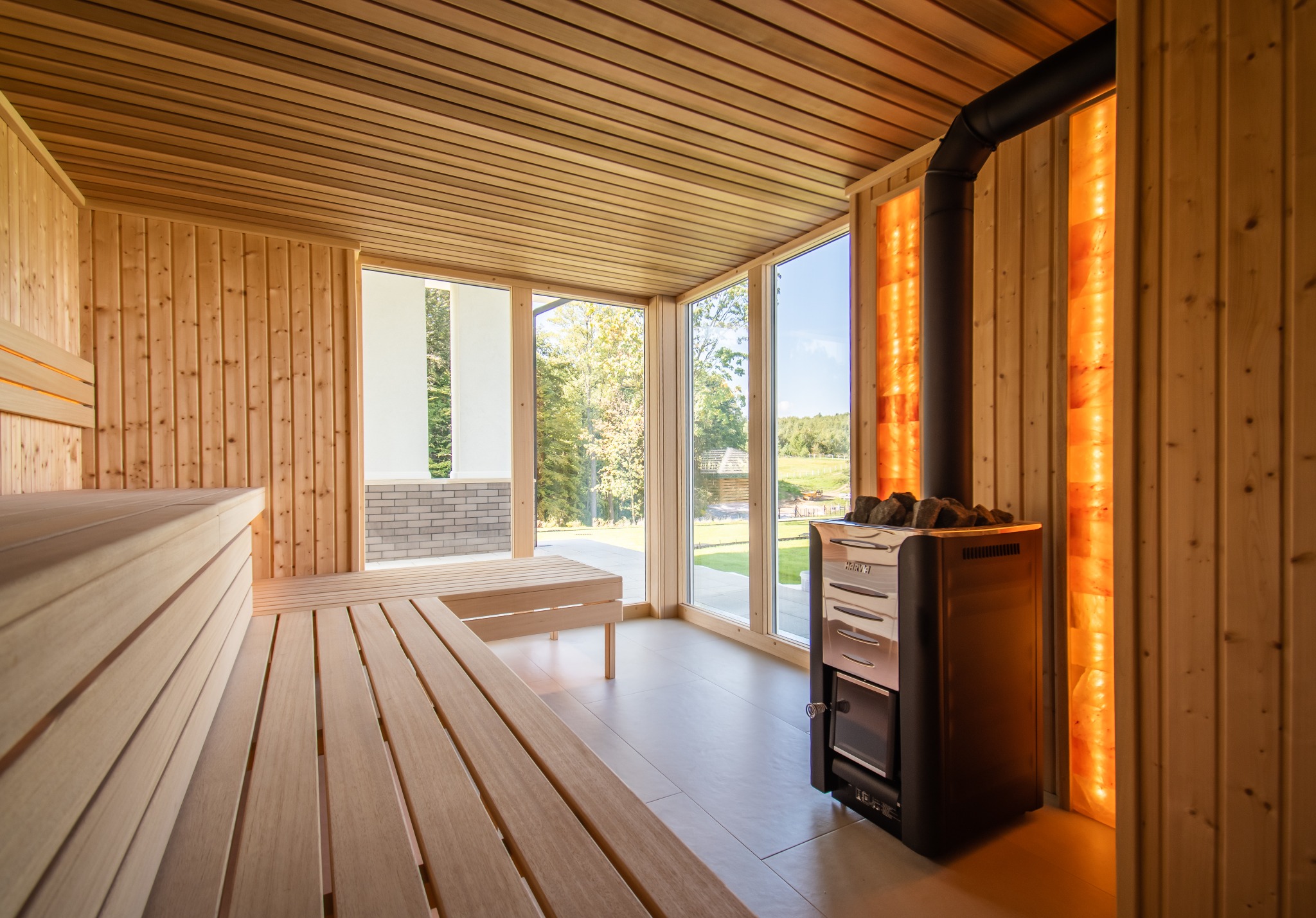 Jasne drewno w saunie z piecem Harvia i oknami z widokiem na zieleń. Podświetlane panele solne dodają ciepła. Minimalistyczny design z ławkami i ceramiczną podłogą.