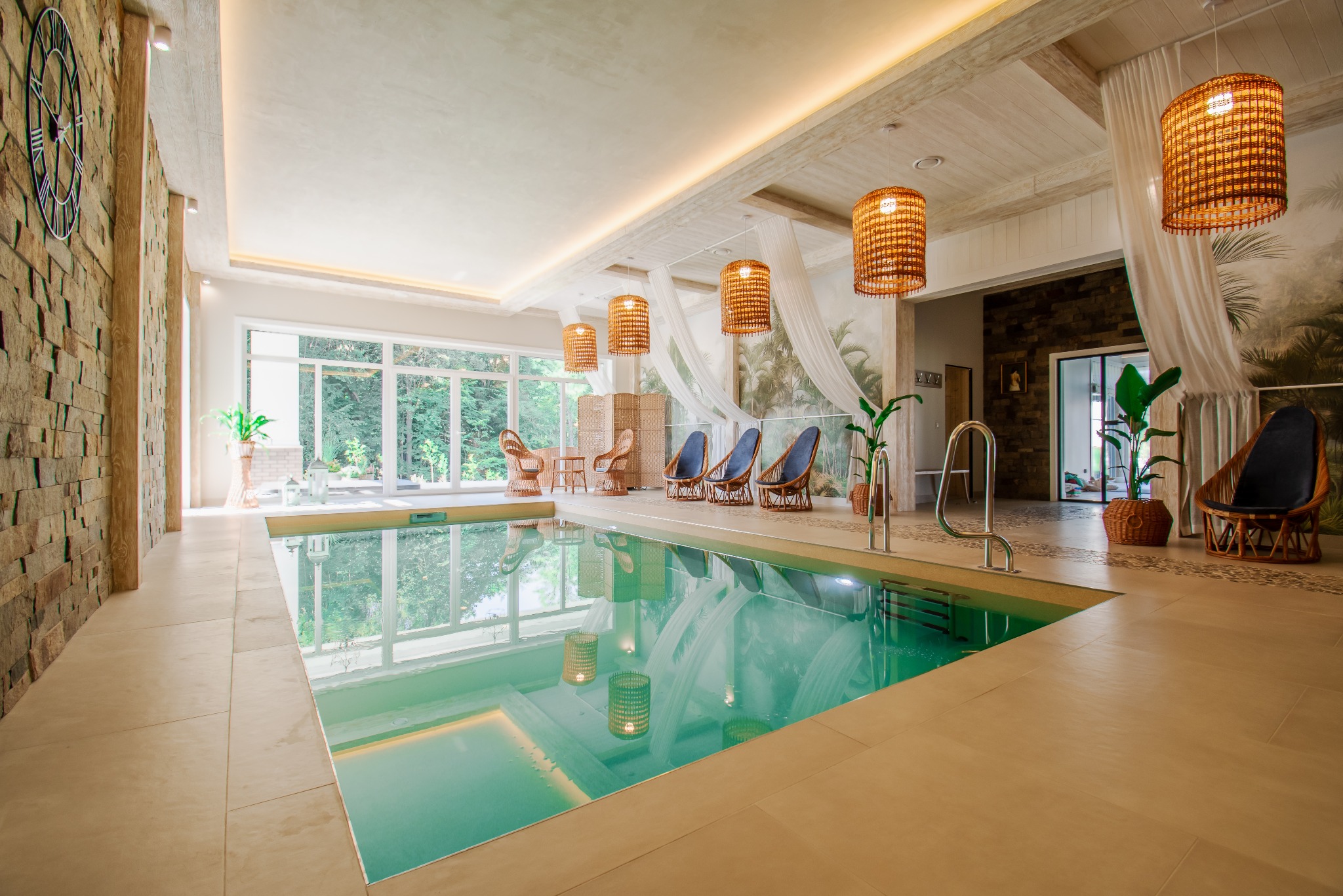 Ekskluzywny kryty basen z turkusową wodą, otoczony jasnymi płytkami i rattanowymi meblami, oświetlony wiszącymi lampami z abażurami. Widok na zieleń za oknem.