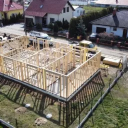Budowa domu szkieletowego w Osiecznej, widok z drona na drewnianą konstrukcję ścian i dachu, w tle zabudowa jednorodzinna i ekipa budowlana przy pracy.