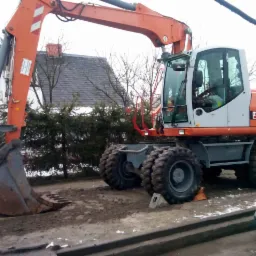 Pomarańczowa koparka kołowa Terex 114C z łyżką, zaparkowana na placu budowy. Widoczne opony z grubym bieżnikiem i kabina operatora. W tle zieleń i fragment dachu budynku.