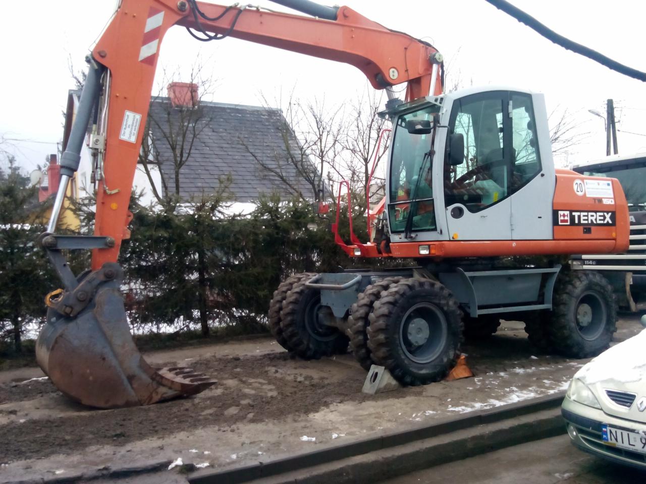 Pomarańczowa koparka kołowa Terex 114C z łyżką, zaparkowana na placu budowy. Widoczne opony z grubym bieżnikiem i kabina operatora. W tle zieleń i fragment dachu budynku.