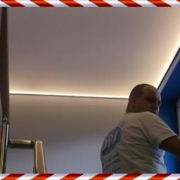 DOMPRO STUDIO Arkadiusz Cybartowicz - Mężczyzna czyści okno w pomieszczeniu z nowoczesnym oświetleniem LED podsufitowym. Widoczny fragment schodów i błękitne ramy okienne.