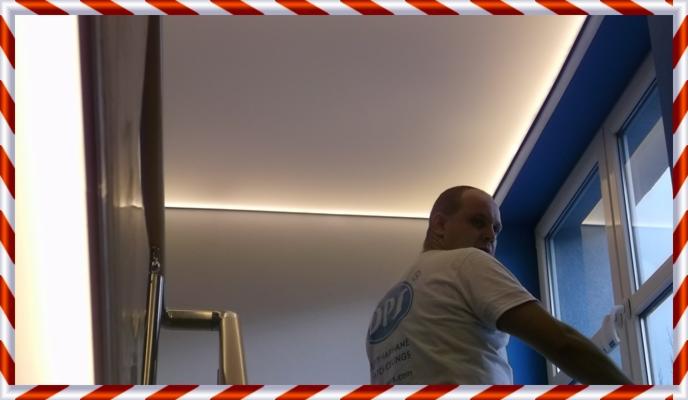 Mężczyzna czyści okno w pomieszczeniu z nowoczesnym oświetleniem LED podsufitowym. Widoczny fragment schodów i błękitne ramy okienne.