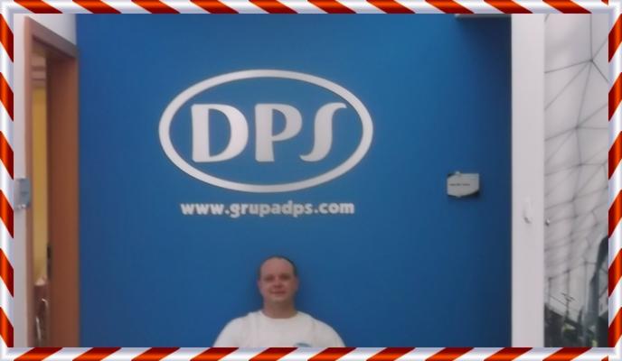 Niebieska ściana biura z logo DPS i adresem www.grupadps.com. Na tle ściany stoi mężczyzna w białej koszulce. Widoczne drzwi i fragment sufitu.