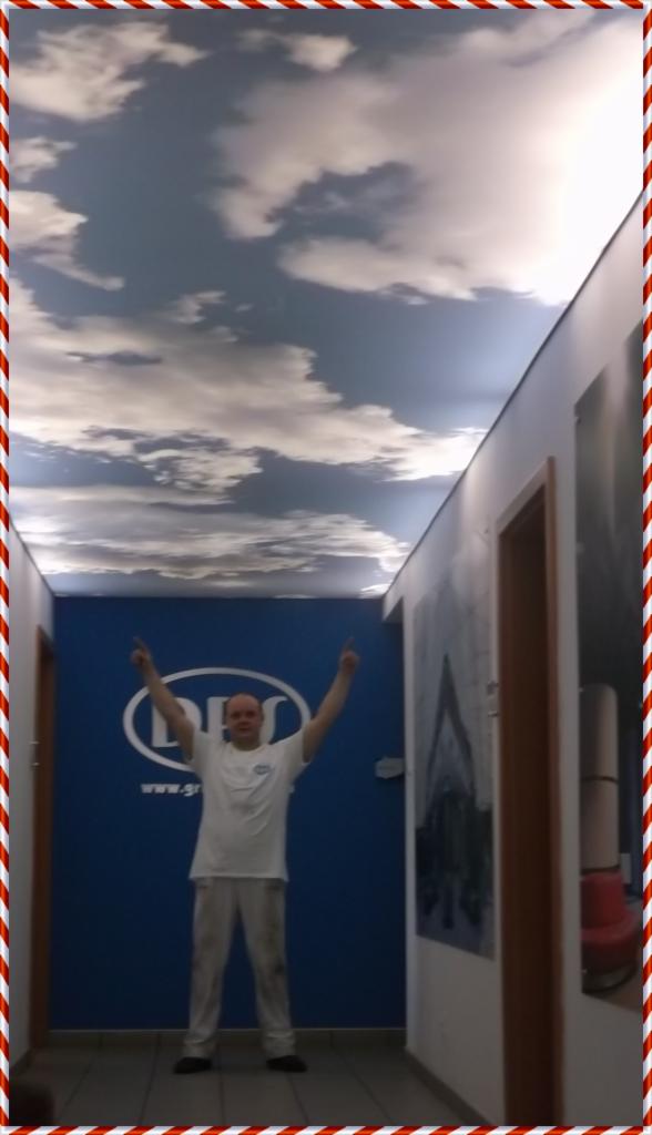 Korytarz biura po remoncie: sufit z efektem nieba, ściany w odcieniach bieli i błękitu. Mężczyzna w stroju roboczym z podniesionymi rękami prezentuje efekt końcowy.