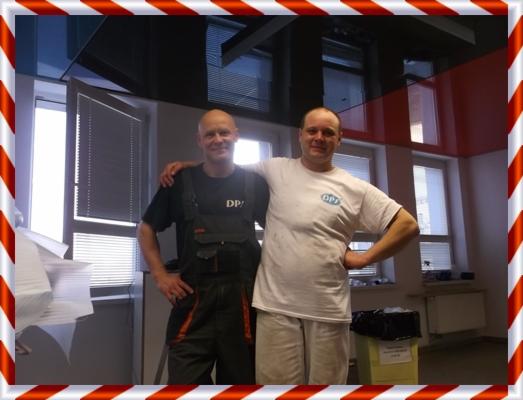 Dwóch mężczyzn w roboczych ubraniach, jeden w kombinezonie, drugi w białej koszulce, stojący w pomieszczeniu z oknami i nowoczesnym, błyszczącym sufitem podwieszanym.