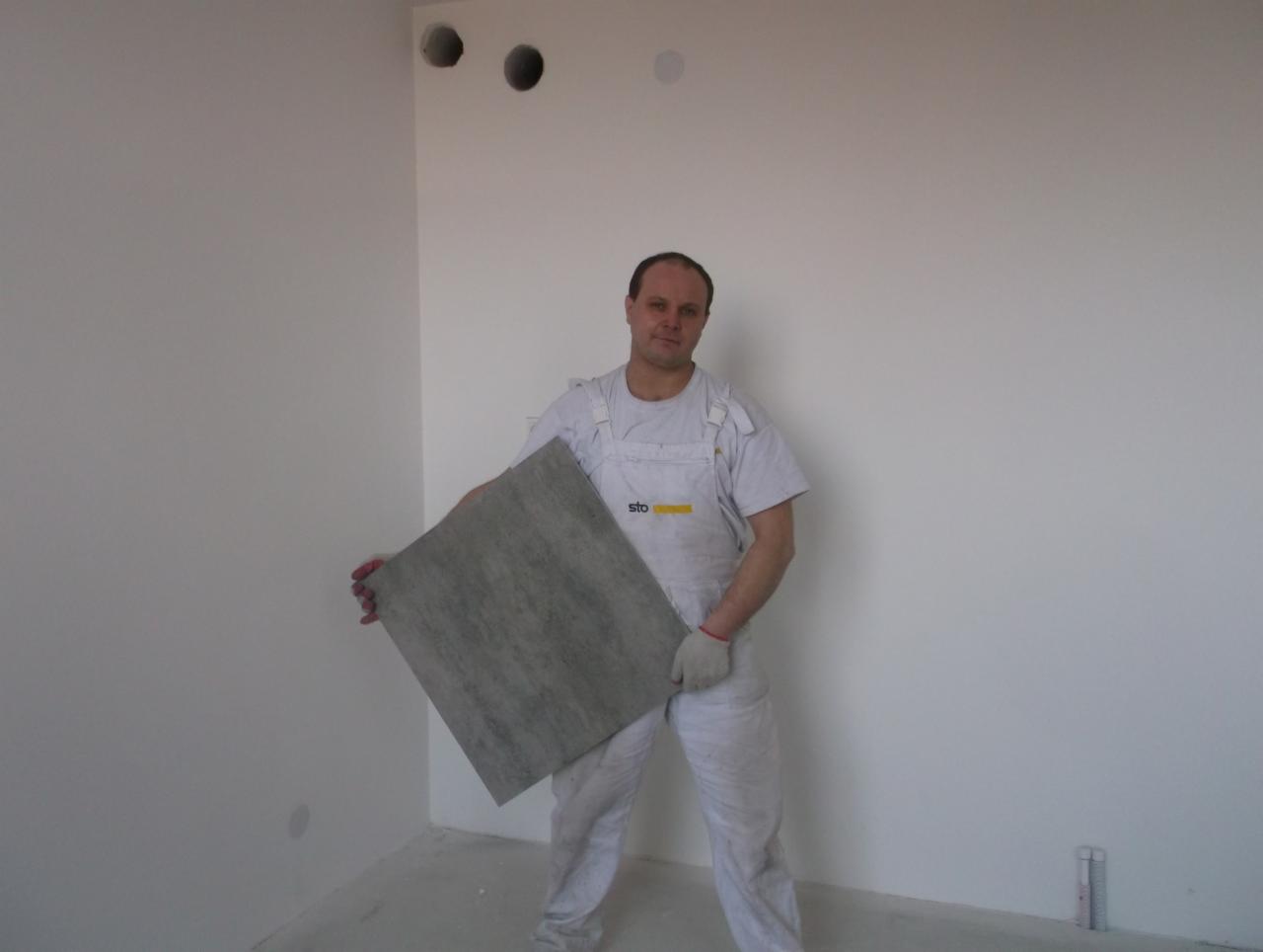 Pracownik w kombinezonie trzyma szarą płytkę na tle białej ściany z otworami. Przygotowanie do remontu, widoczne rękawice i logo firmy STO na ubraniu.