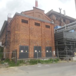 PlaniProjekt.pl - Zewnętrzny widok ceglanego budynku przemysłowego z trzema szarymi drzwiami z ostrzeżeniami, metalowymi rurami i rusztowaniami, częściowo ogrodzony siatką.