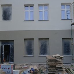 Goryl Mateusz usługi budowlane - Dwukolorowa elewacja budynku w trakcie prac. Okna zabezpieczone folią, widoczne materiały budowlane i pracownik w trakcie przygotowań. Częściowo widoczne rusztowanie.