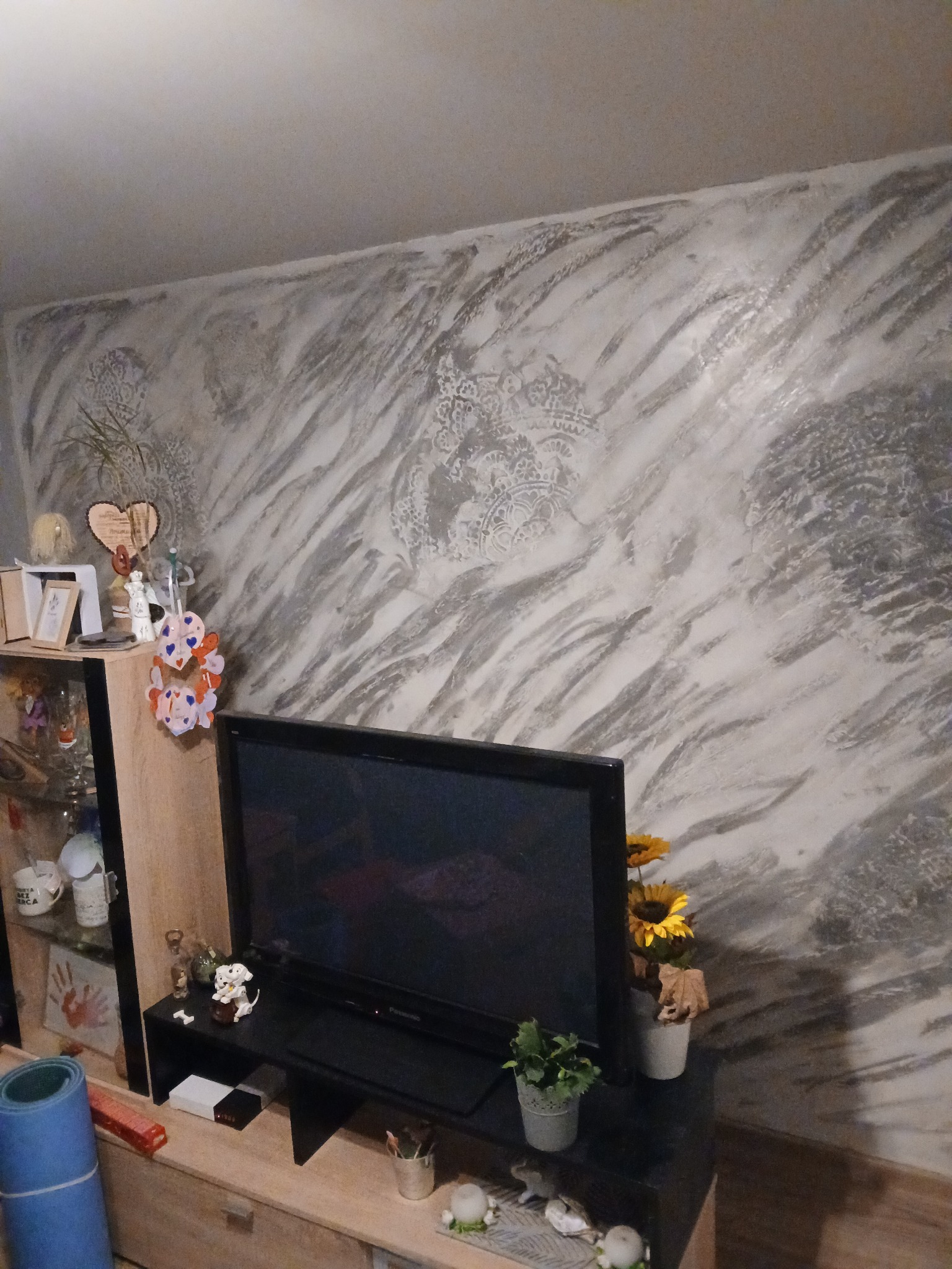 Ściana w salonie z dekoracyjnym, marmurkowym efektem tynku w odcieniach szarości i bieli, widoczna za telewizorem i elementami dekoracyjnymi na szafce RTV.