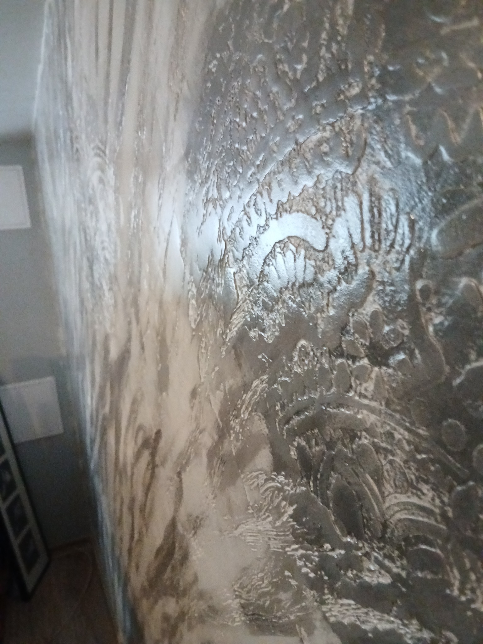 Dekoracyjna ściana wewnętrzna z efektem strukturalnym w odcieniach szarości i bieli, widoczna pod kątem, z elementami wystroju w tle.