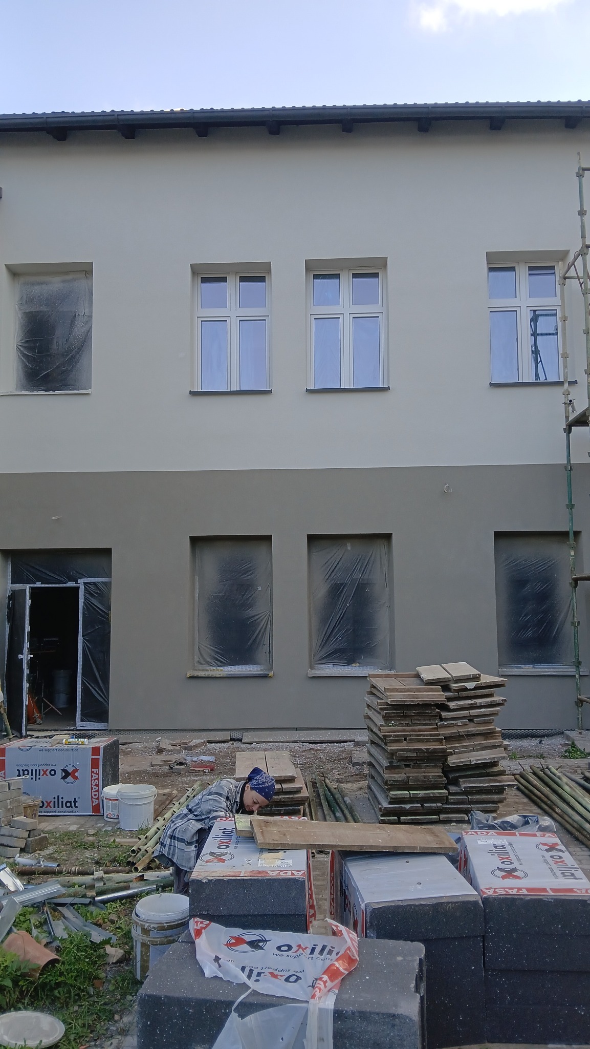 Dwukolorowa elewacja budynku w trakcie prac. Okna zabezpieczone folią, widoczne materiały budowlane i pracownik w trakcie przygotowań. Częściowo widoczne rusztowanie.