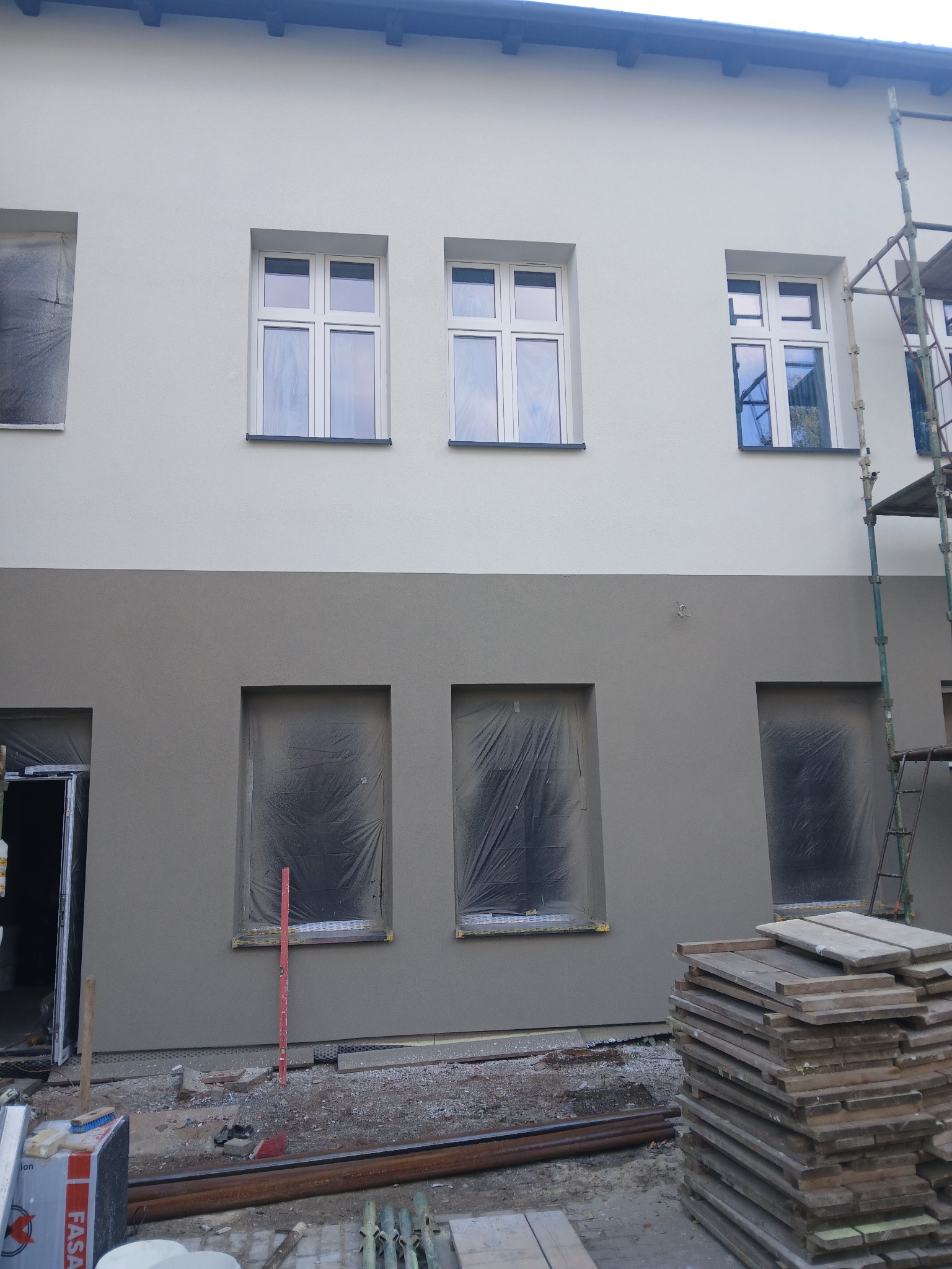 Dwukolorowa elewacja budynku w trakcie prac wykończeniowych. Okna zabezpieczone folią, widoczne rusztowania i materiały budowlane.