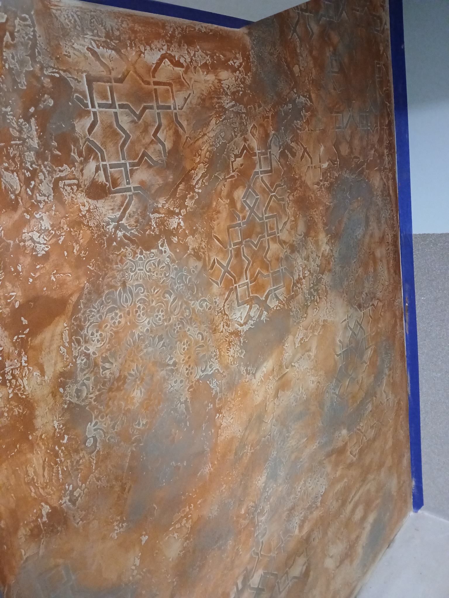 Dekoracyjna ściana w odcieniach brązu i szarości z geometrycznymi i kwiatowymi wzorami, zabezpieczona niebieską taśmą malarską przy suficie i ścianie bocznej.