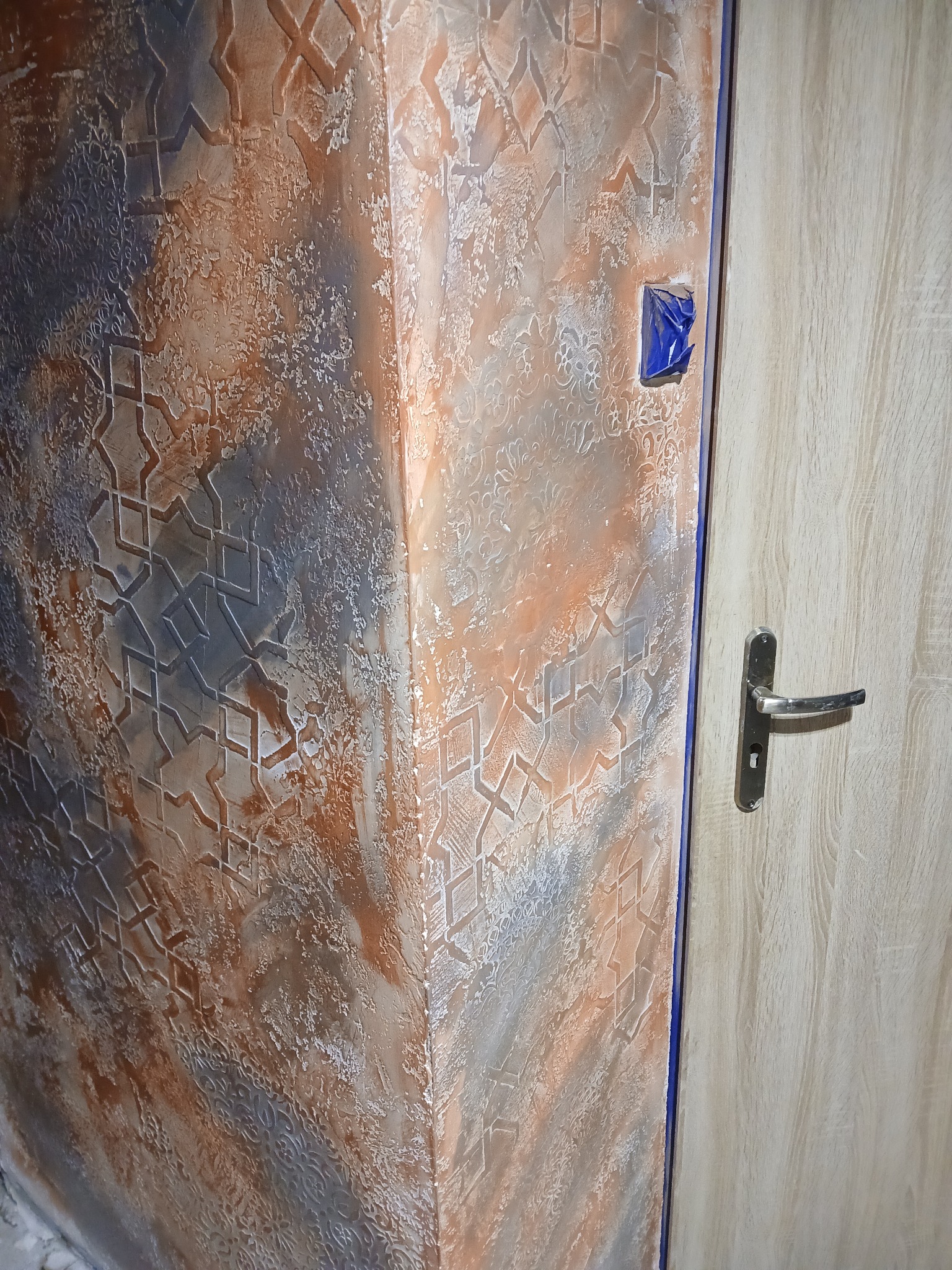 Dekoracyjna ściana w odcieniach brązu i szarości z geometrycznym wzorem, sąsiadująca z jasnymi drzwiami. Fragment zabezpieczonego włącznika.