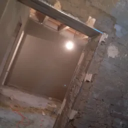 Surowy stan wnętrza w trakcie remontu: świeżo otynkowane ściany z widoczną metalową konstrukcją nad otworem drzwiowym, narzędzia i przewody na podłodze.