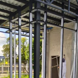 Pracownik w białym kombinezonie wyrównuje mokrą ścianę w surowym stanie, widoczna metalowa konstrukcja budynku i elementy instalacji.