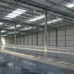 Wnętrze hali przemysłowej z betonową posadzką, widoczne stalowe słupy konstrukcyjne i świetliki w dachu, ślady po pracach budowlanych na podłodze.