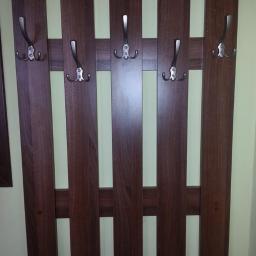 Robert Urban Zakład Produkcyjno Usługowy URBAN meble - Wieszak ścienny z ciemnego drewna z pięcioma metalowymi haczykami, zamontowany na jasnej ścianie. Widoczne detale łączeń elementów drewnianych.