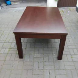 Masywny, prostokątny stół z ciemnego drewna, ustawiony na brukowanym podwórku, z widocznym warsztatem w tle. Minimalistyczny design, solidne nogi.