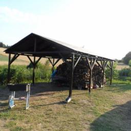 Robert Urban Zakład Produkcyjno Usługowy URBAN meble - Drewniana wiata z ciemnym dachem, wypełniona stosem drewna. Na trawie grill i fragment samochodu. W tle pola i zieleń. Ujęcie w letnim słońcu.