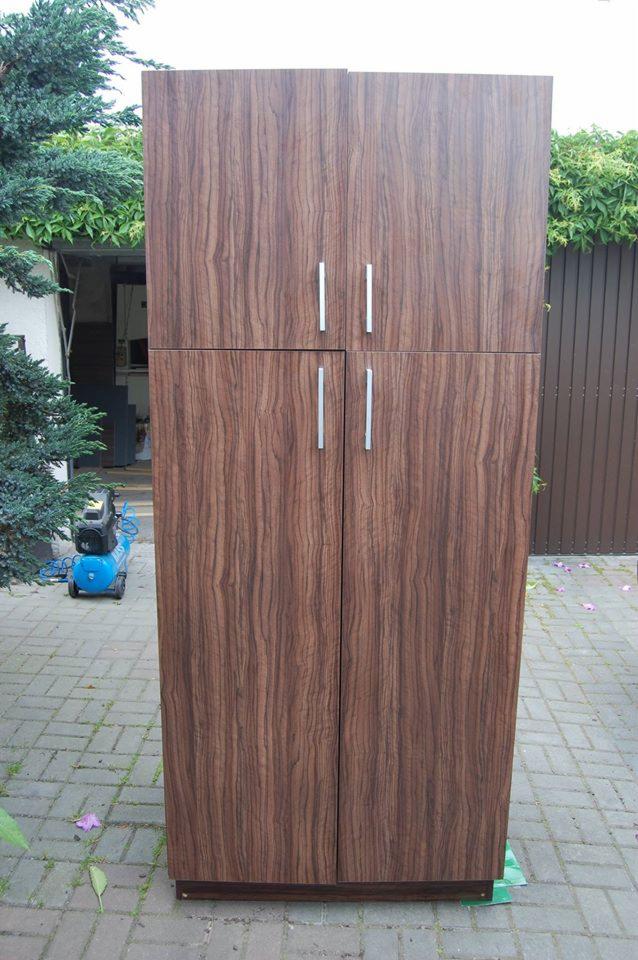 Wysoka, brązowa szafa z czterema drzwiami i długimi, srebrnymi uchwytami, ustawiona na kostce brukowej, z zielenią w tle.