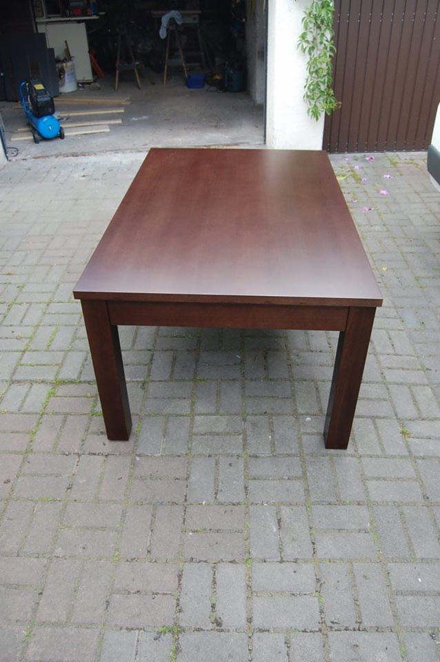 Masywny, prostokątny stół z ciemnego drewna, ustawiony na brukowanym podwórku, z widocznym warsztatem w tle. Minimalistyczny design, solidne nogi.