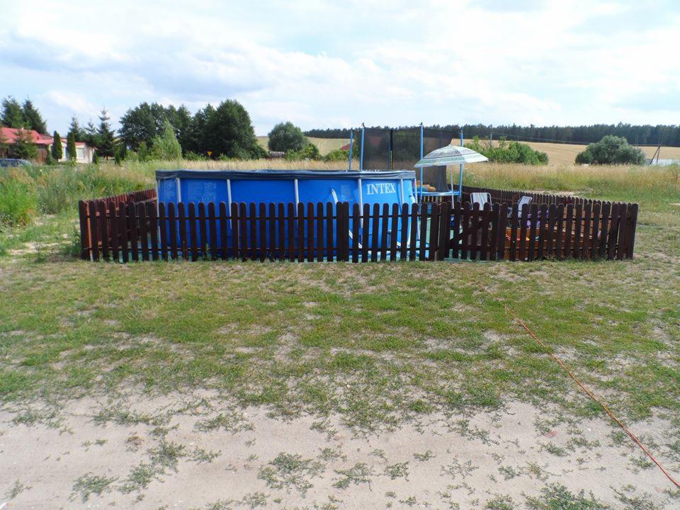 Ogrodzony, naziemny basen Intex na trawiastym podwórku z trampoliną w tle, otoczony niskim, drewnianym płotem sztachetowym. W tle drzewa i pola.