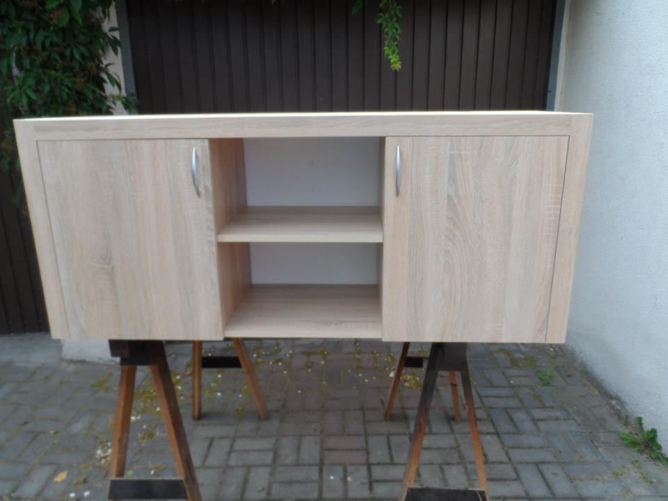 Jasna komoda z drewnopodobnym dekorem, zamykana, z półkami w środku. Mebel stoi na kozłach, w Szamocinie. Widok z przodu, na zewnątrz.