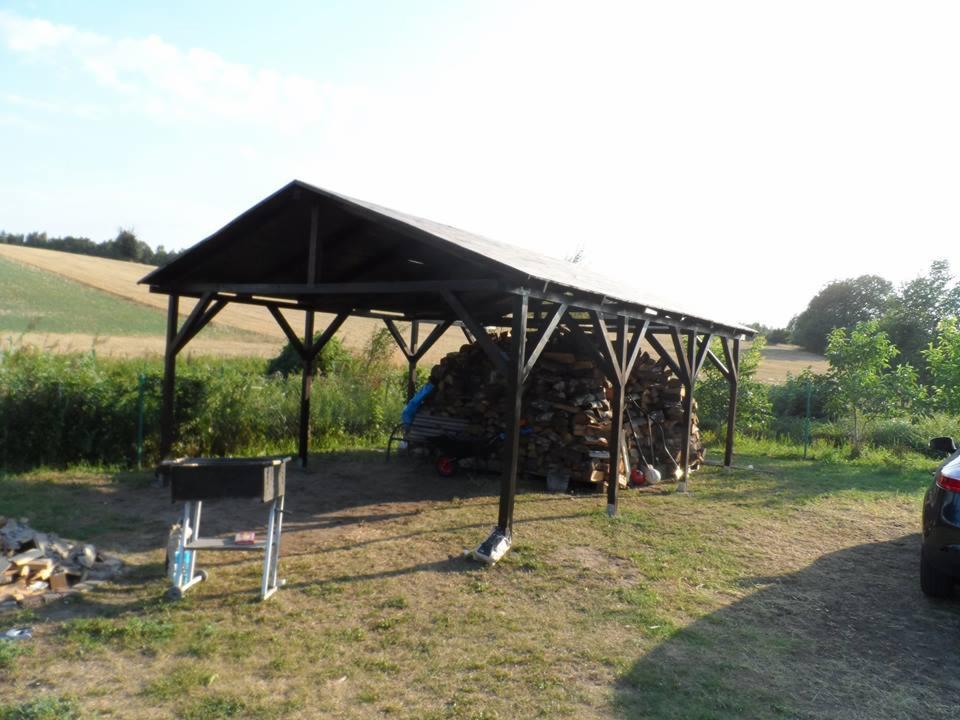 Drewniana wiata z ciemnym dachem, wypełniona stosem drewna. Na trawie grill i fragment samochodu. W tle pola i zieleń. Ujęcie w letnim słońcu.