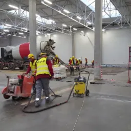 Prace w hali produkcyjnej: ekipa w żółtych kamizelkach obrabia wylewkę przy użyciu frezarki do betonu i odkurzacza przemysłowego, w tle betoniarka i zapasy materiałów.