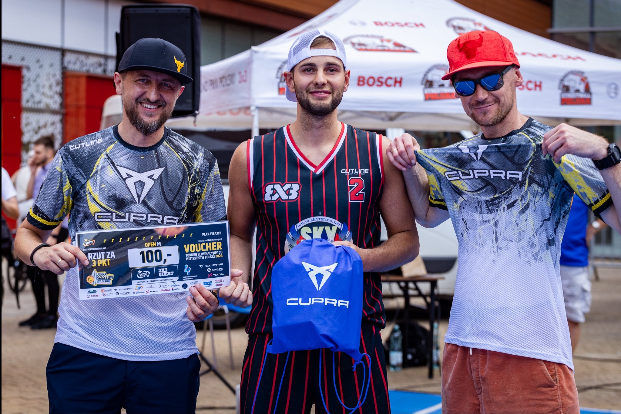 Trzy osoby prezentują koszulki i torbę z logo Cupra na tle namiotu reklamowego podczas turnieju koszykówki 3x3.
