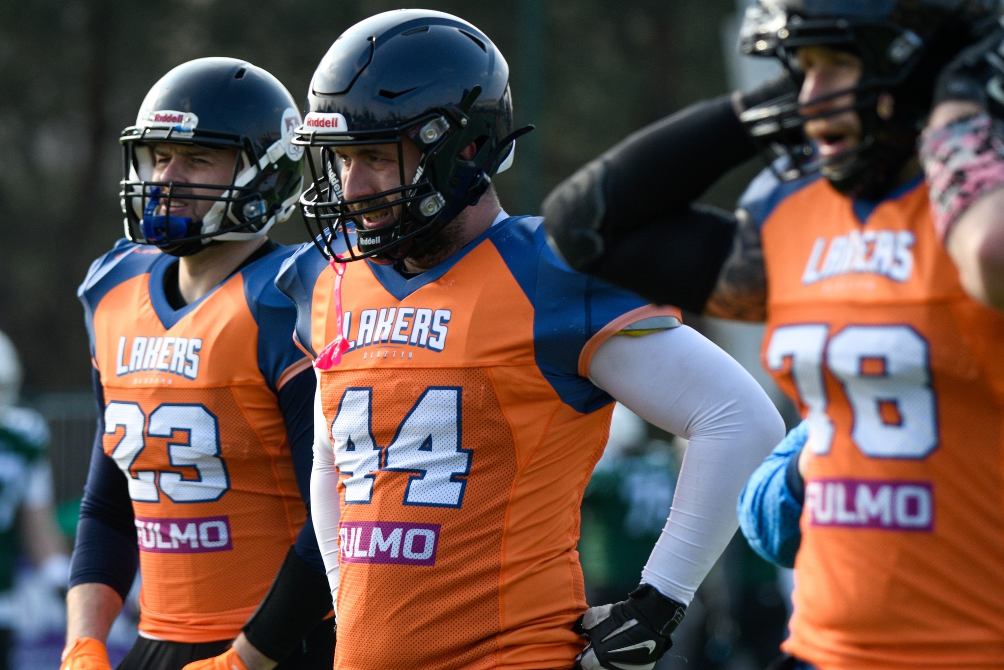 Zawodnicy futbolu amerykańskiego w pomarańczowo-niebieskich strojach z numerami i nazwą drużyny 'Lakers Olsztyn'. Kaski, ochraniacze, rękawice.