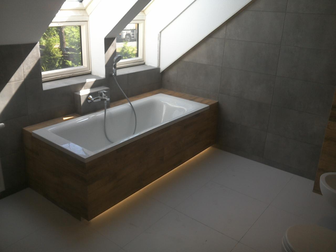 Nowoczesna łazienka na poddaszu z wanną obudowaną drewnem i podświetleniem LED. Szare płytki na ścianach, biała podłoga i okna dachowe.