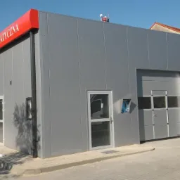 Nowoczesna, szara, bezobsługowa myjnia samochodowa z czerwonym daszkiem i napisem, widok z zewnątrz w słoneczny dzień.