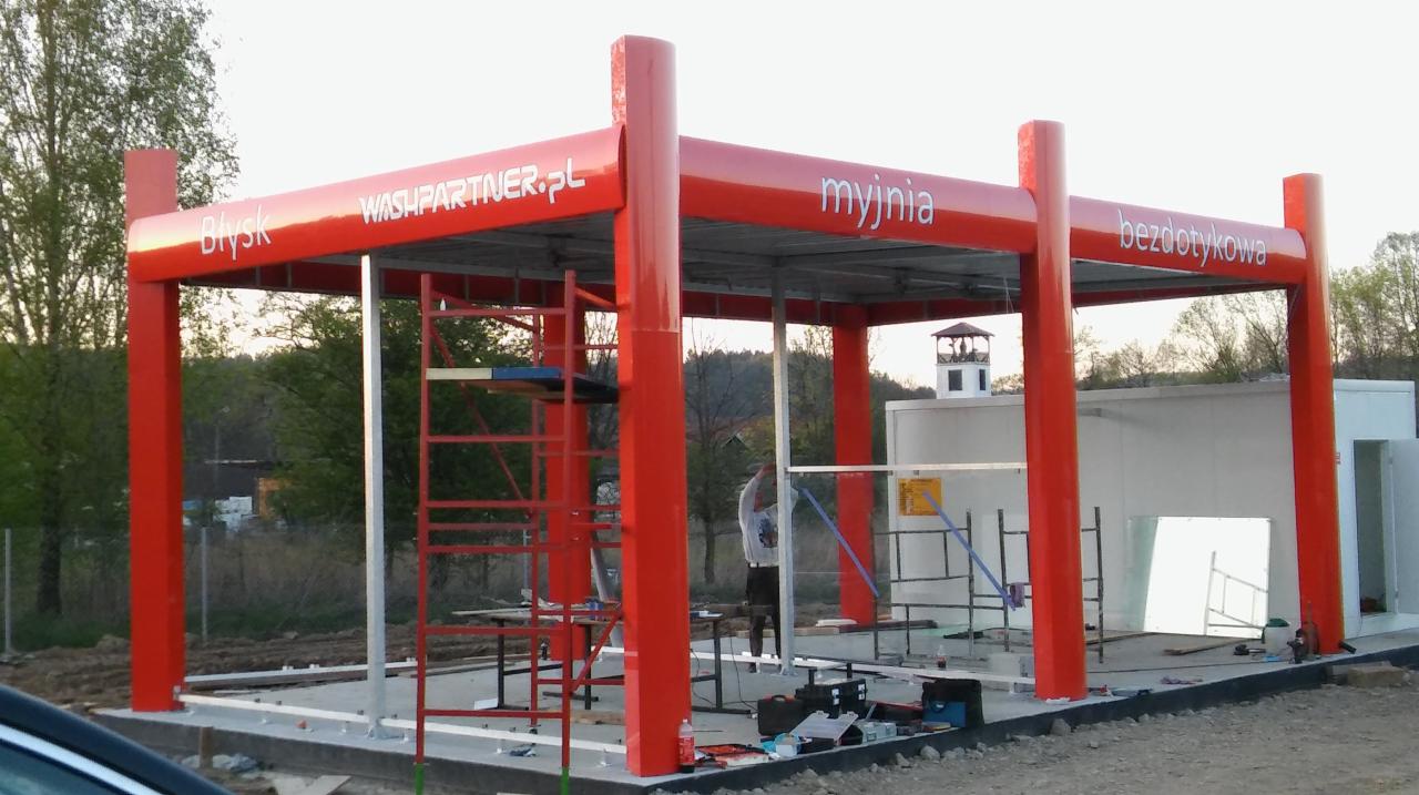 Budowa bezdotykowej myjni samochodowej z czerwonymi elementami konstrukcyjnymi i napisem 'Błysk WASHPARTNER.pl', 'myjnia', 'bezdotykowa', widoczne rusztowania i pracownik w trakcie montażu.
