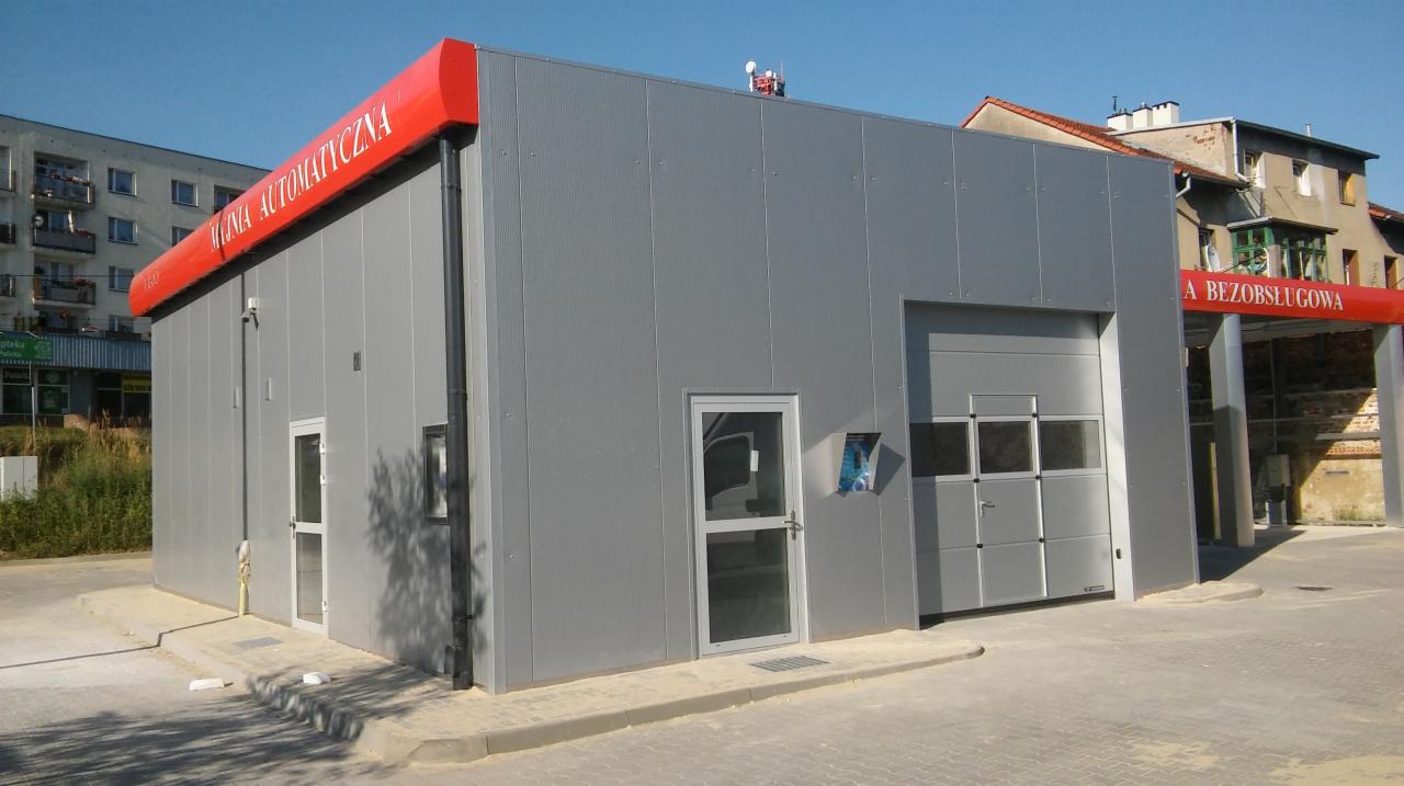 Nowoczesna, szara, bezobsługowa myjnia samochodowa z czerwonym daszkiem i napisem, widok z zewnątrz w słoneczny dzień.