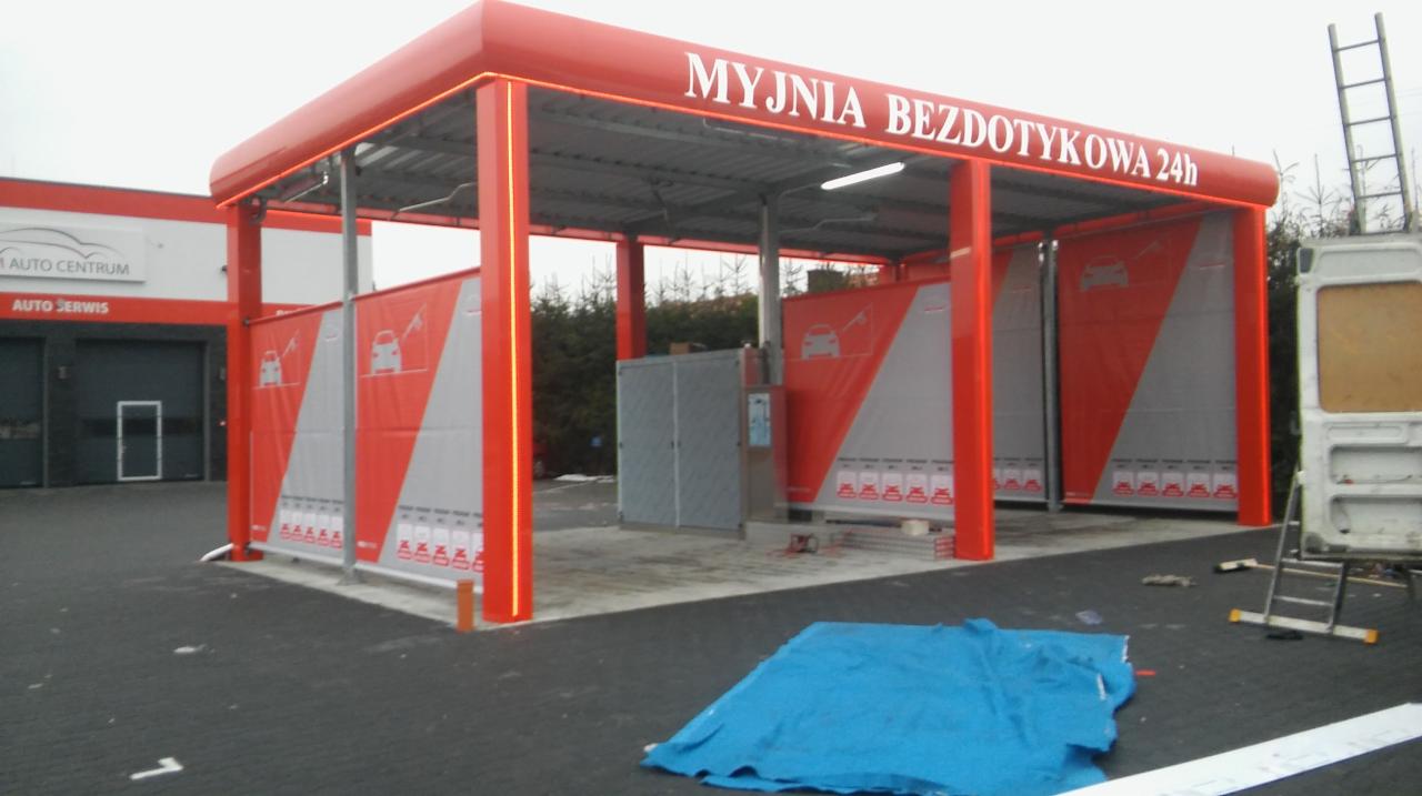 Zewnętrzny widok stanowisk myjni bezdotykowej 24h z czerwonymi elementami konstrukcyjnymi i panelami z grafiką samochodów, częściowo zasłoniętej niebieską plandeką i z drabiną opartą o samochód...