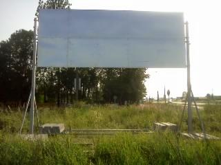 Pusta, metalowa tablica reklamowa na konstrukcji wsporczej, widoczna z perspektywy trawy, z drzewami i drogą w tle.