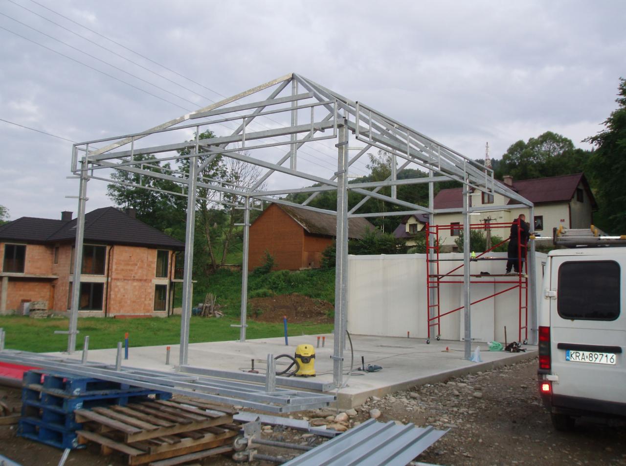 Montaż szkieletu stalowego konstrukcji w trakcie budowy, widoczny pracownik na rusztowaniu, części konstrukcji na paletach, budynki w tle.