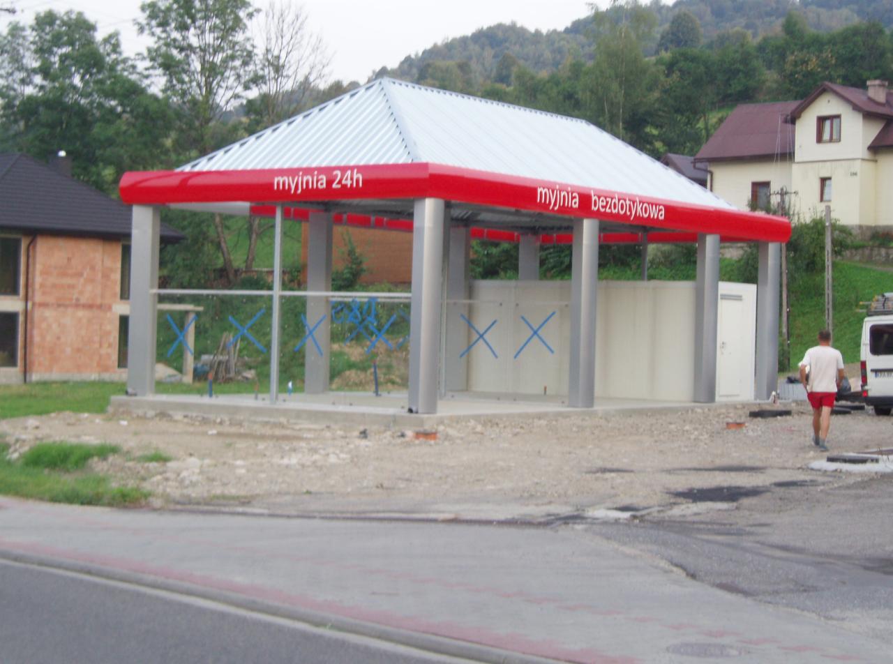 Nowa, bezdotykowa myjnia 24h z czerwonymi elementami konstrukcyjnymi i srebrnym dachem w trakcie budowy, widoczne zabezpieczenia niebieską taśmą, w tle budynki mieszkalne i zalesione wzgórza.
