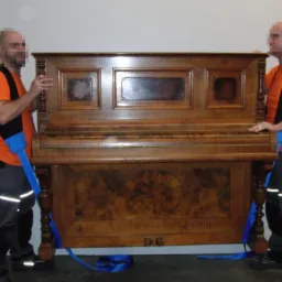 Dwóch mężczyzn w pomarańczowych koszulkach transportuje pianino za pomocą specjalnych pasów. Widoczne detale drewnianej obudowy instrumentu i elementy stroju robotników.