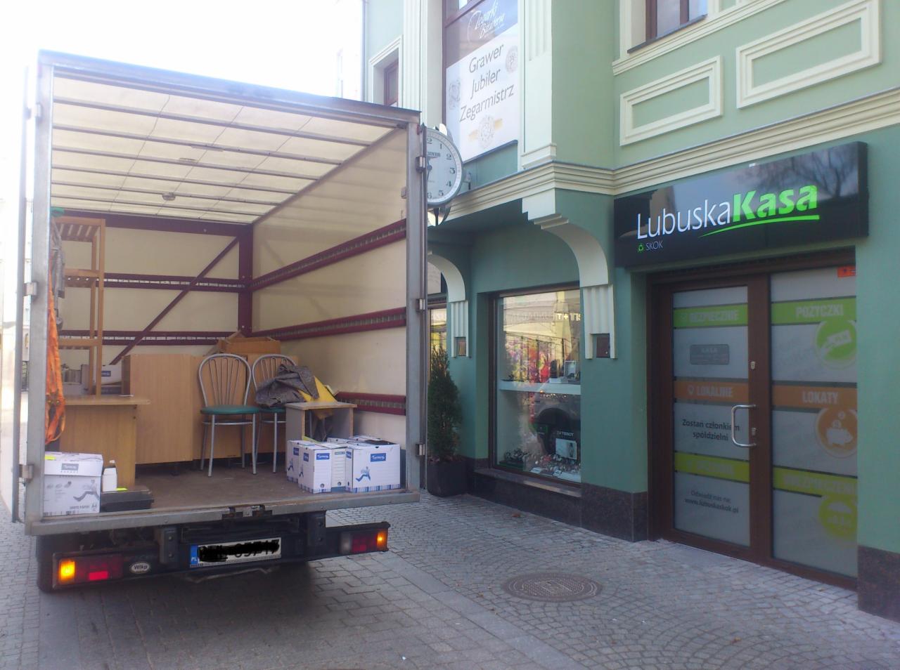 Otwarta paka busa z meblami w trakcie przeprowadzki w Zielonej Górze. Widoczne biurko, krzesła i kartony. W tle budynek z szyldem Lubuska Kasa.