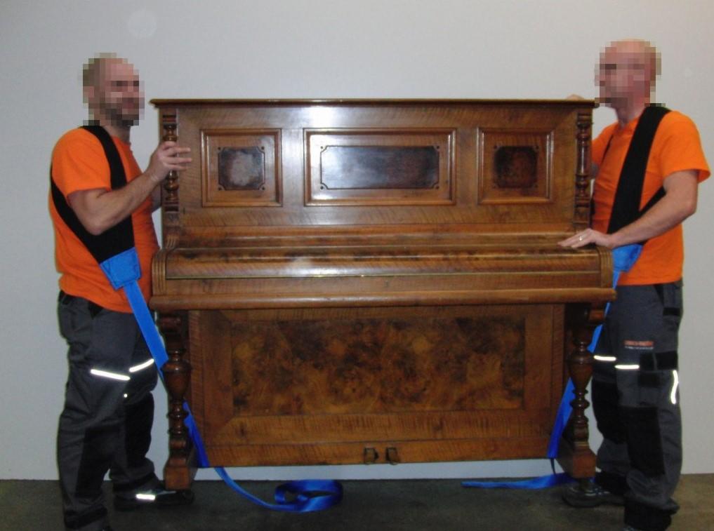 Dwóch mężczyzn w pomarańczowych koszulkach transportuje pianino za pomocą specjalnych pasów. Widoczne detale drewnianej obudowy instrumentu i elementy stroju robotników.